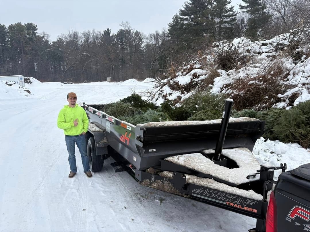 Person in bright yellow jacket beside a trailer loaded with Christmas trees on a snowy road.