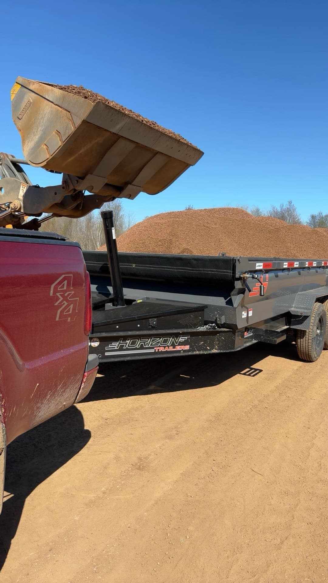Truck loading soil onto a black trailer with a pile of material in the background on a sunny day.