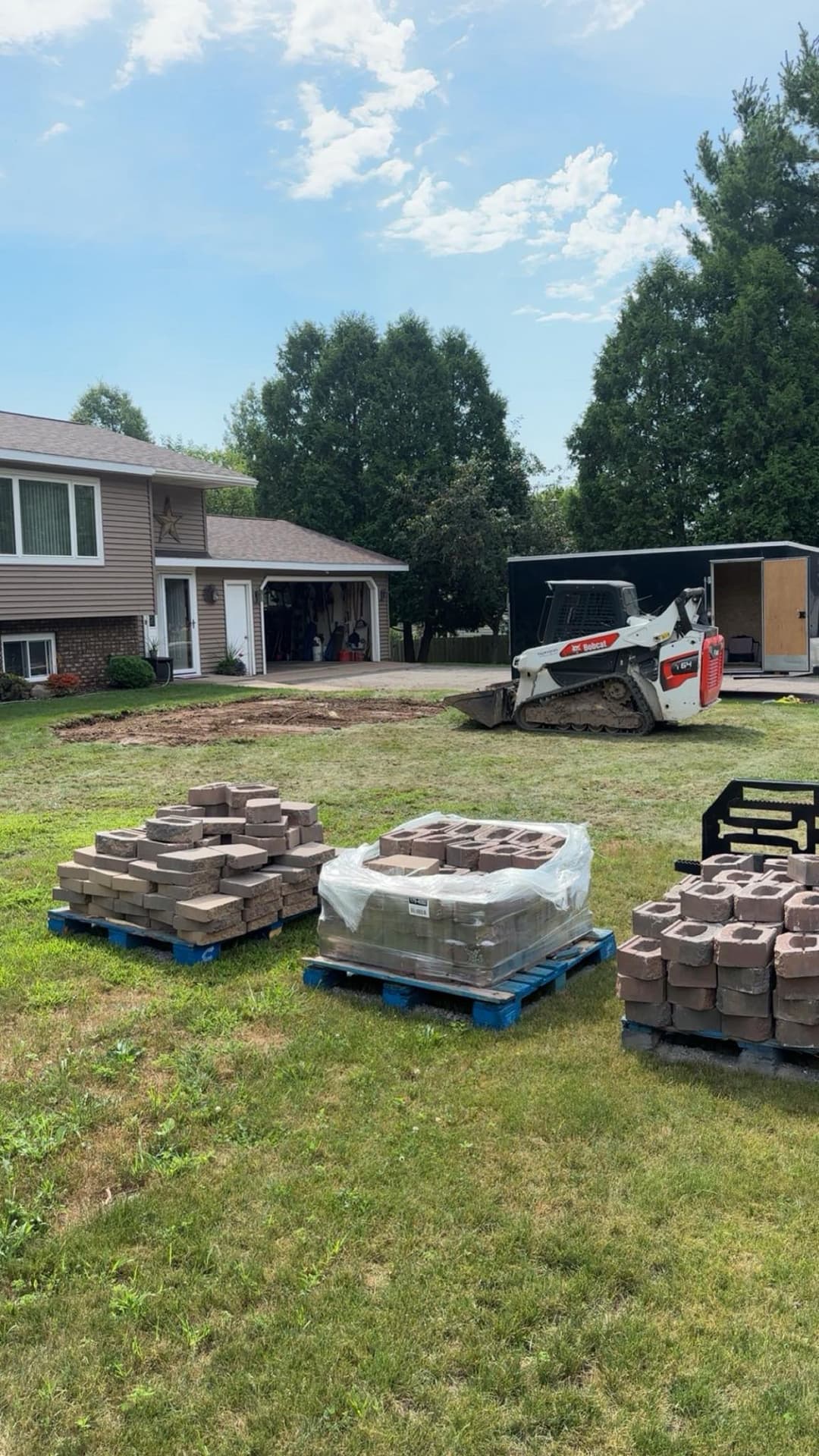 Pallets of paving stones in a yard with a bobcat and a house in the background.
