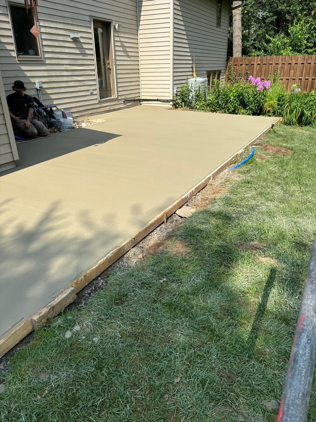 Freshly poured concrete patio beside a house, with green grass and garden in view.