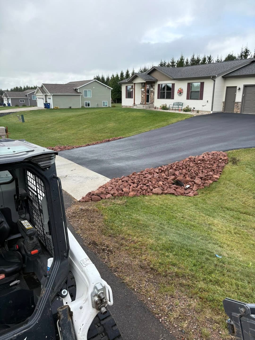 Bobcat loader near freshly laid asphalt driveway with red rock border in residential area.