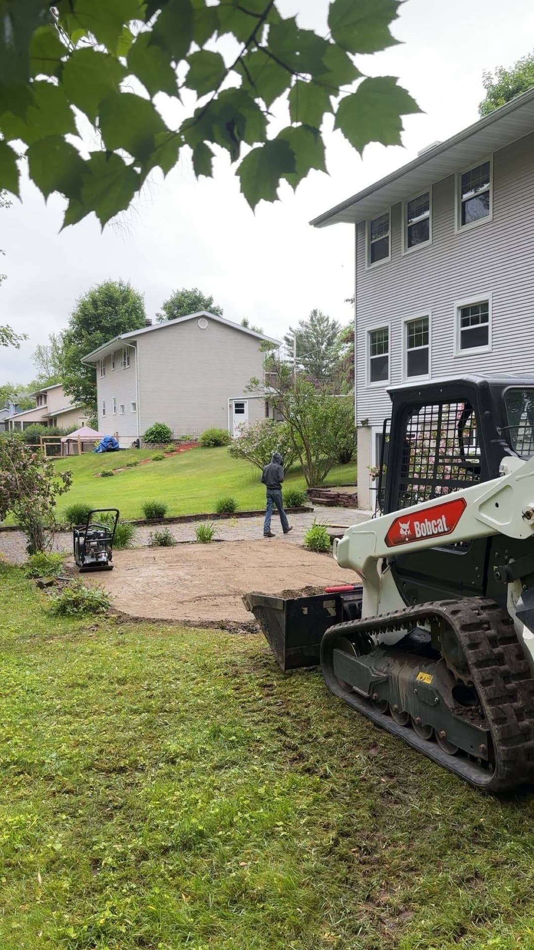 Bobcat machinery landscaping a yard with a worker planting greenery near a house.