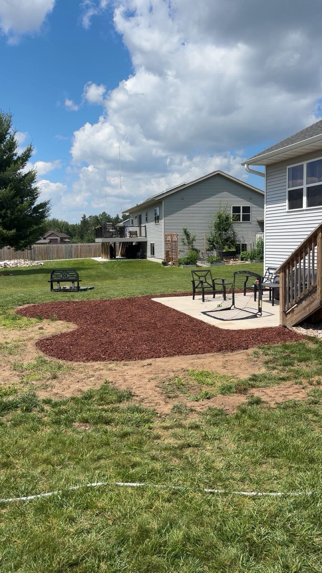 Backyard scene featuring a mulched area, patio with chairs, and nearby homes under a cloudy sky.