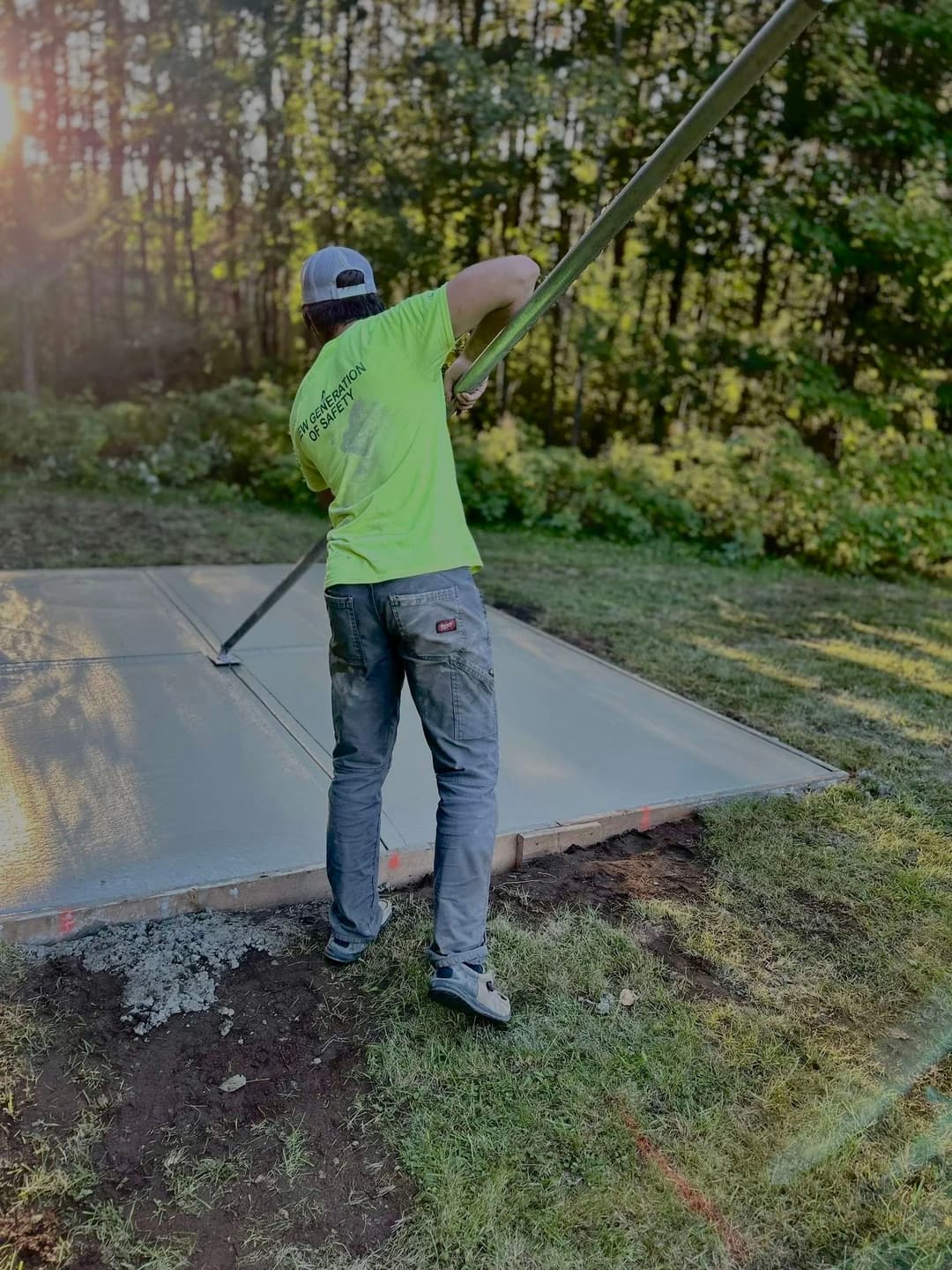 Worker leveling concrete slab outdoors in sunlight, wearing bright green shirt and gray pants.