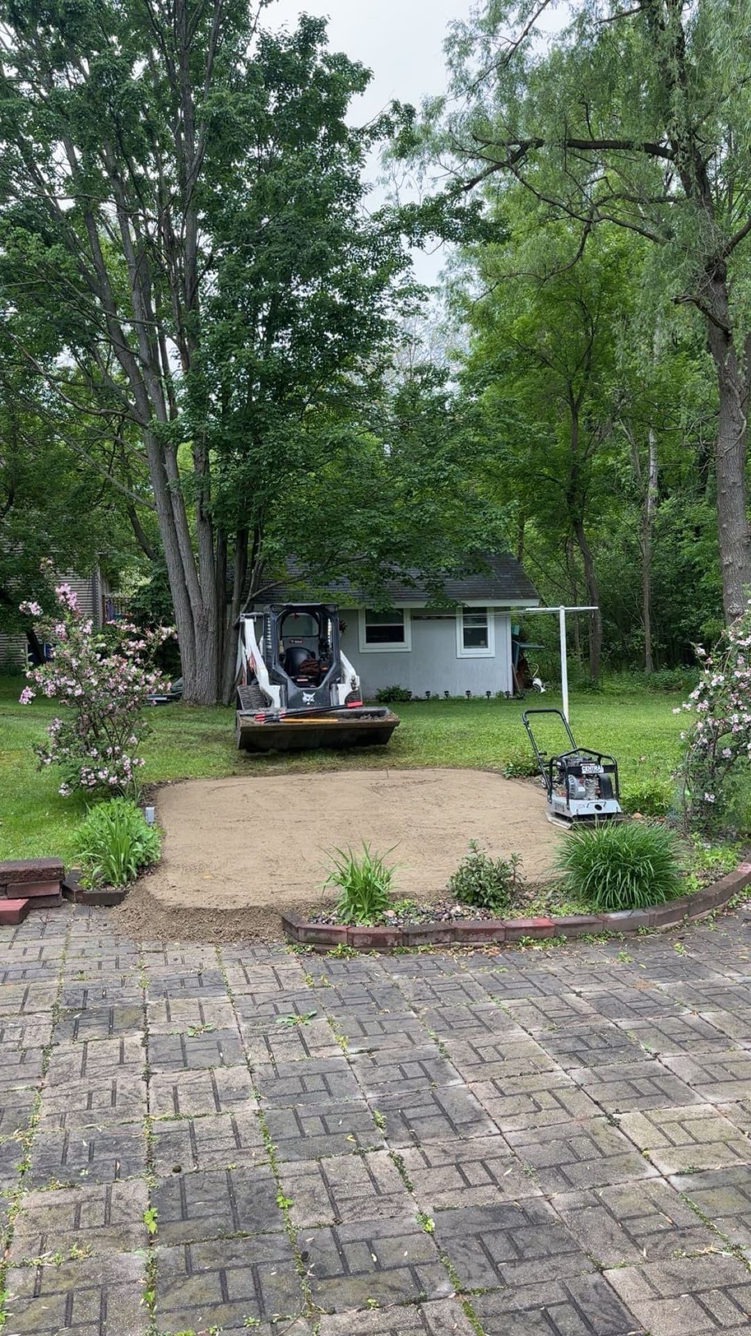 Bobcat loader on a landscaped yard with trees and a small house in the background.