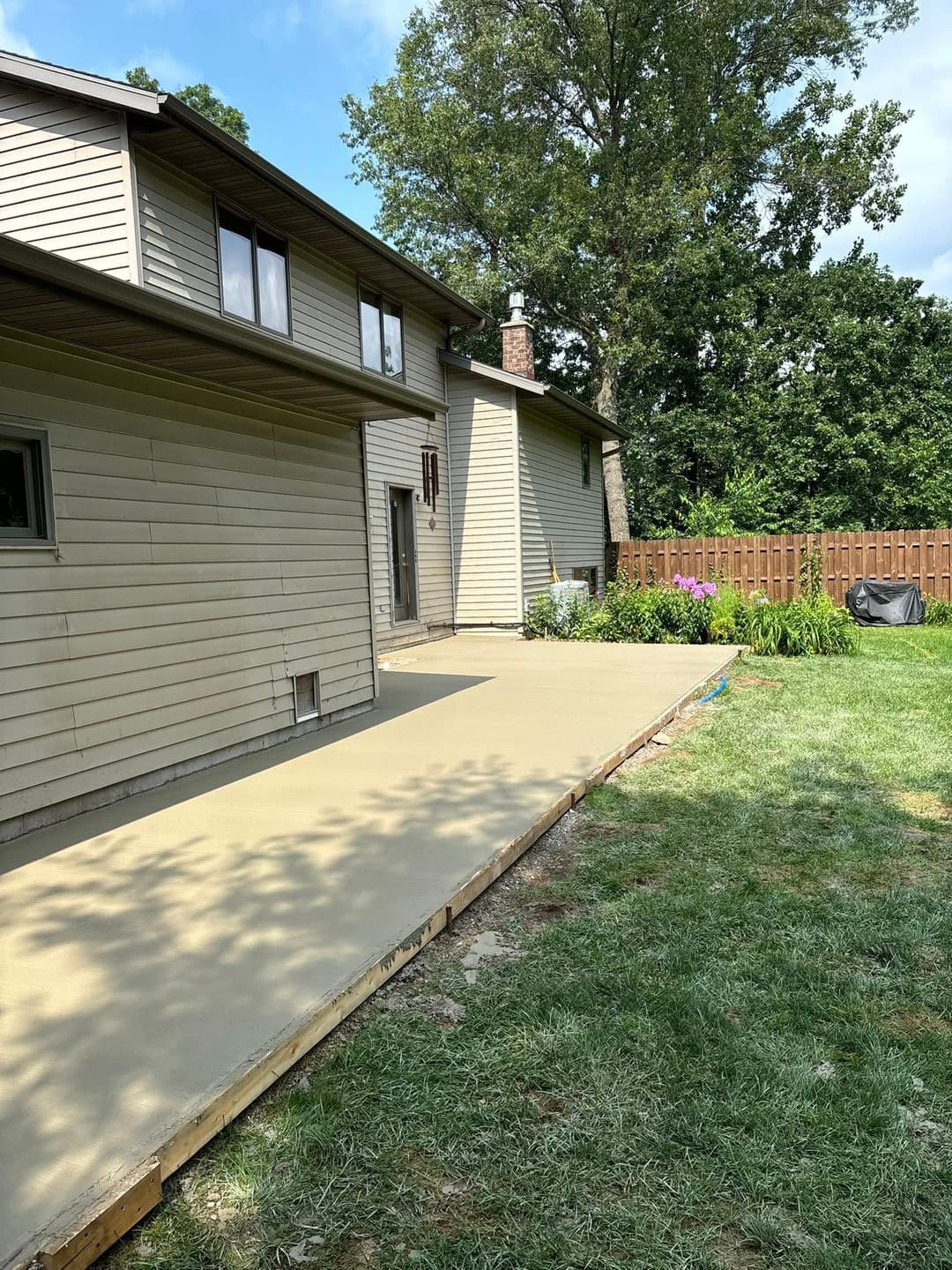 Concrete patio installation beside a house in a landscaped backyard with grass and plants.