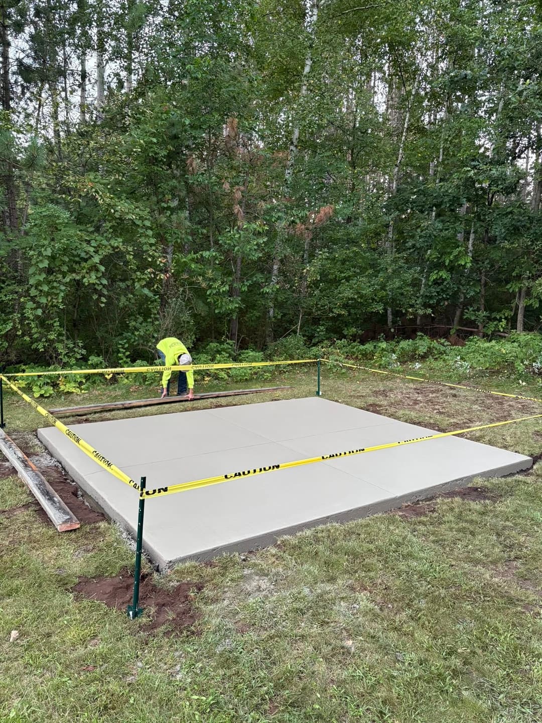 Worker finishing a freshly poured concrete slab, surrounded by caution tape in a wooded area.