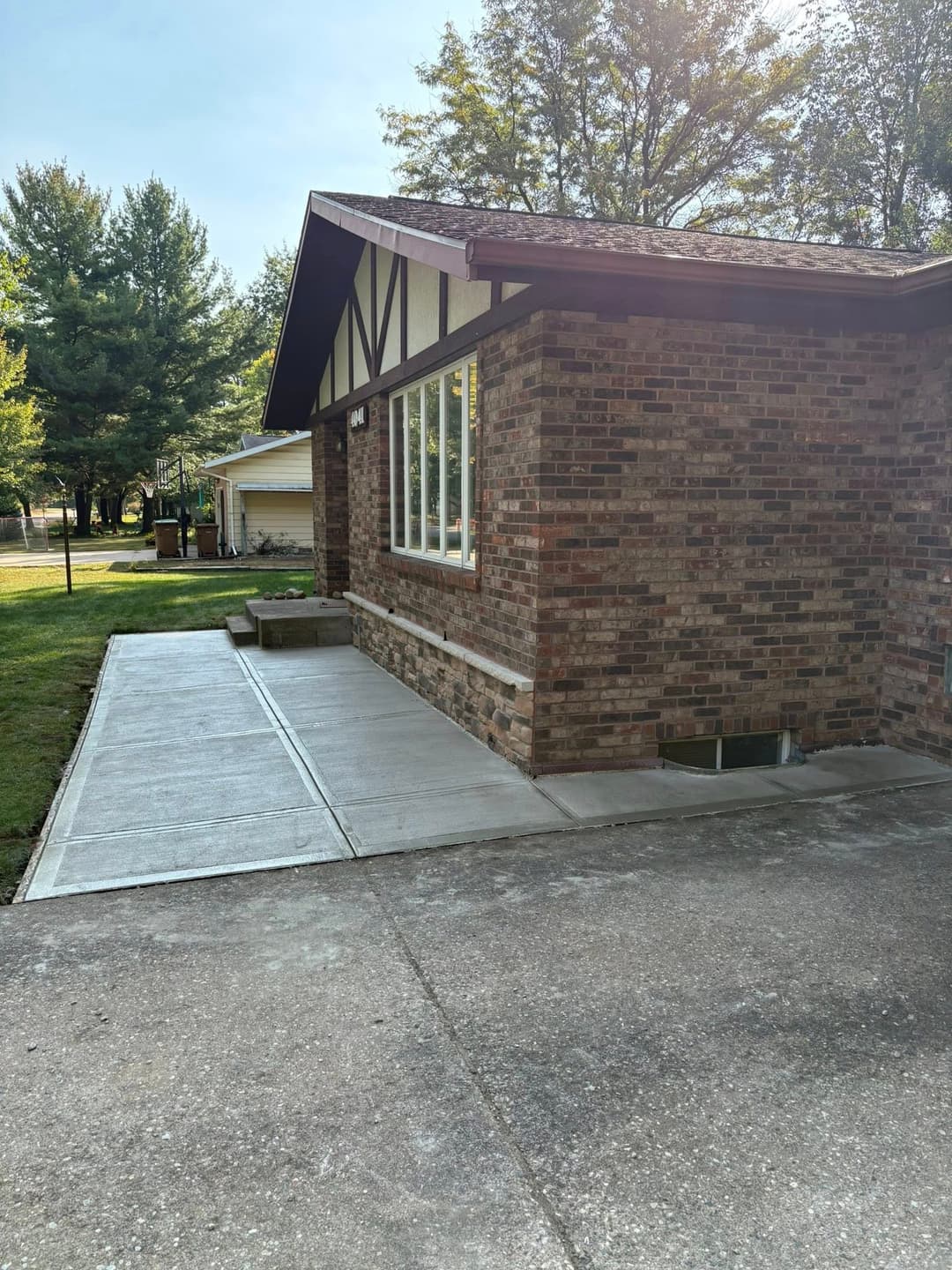 Brick house corner with large windows and freshly poured concrete walkway. Lush green yard.