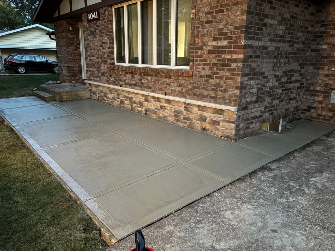 Freshly poured concrete walkway adjacent to a brick house with a lawn and driveway.