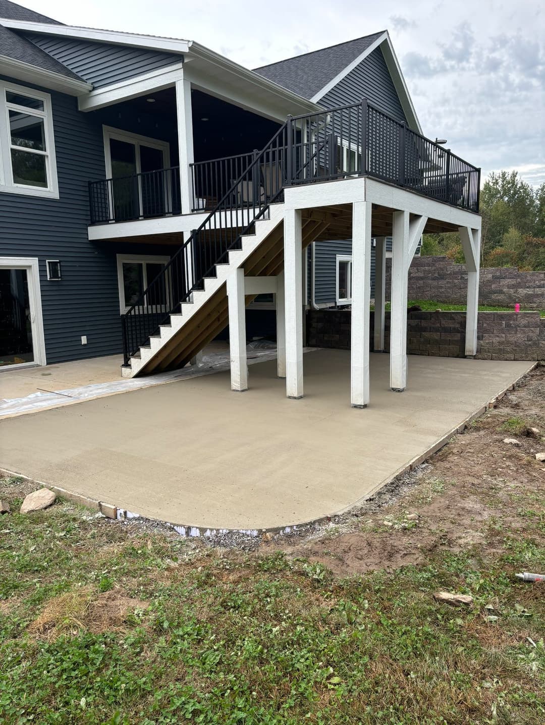 Newly poured concrete patio beneath a raised deck with black railings and modern home exterior.