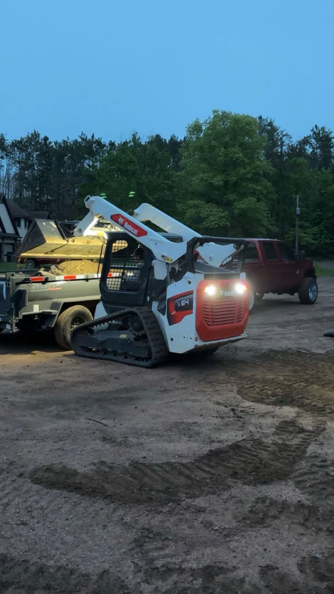 Bobcat E164 mini skid steer parked at dusk near a trailer and pickup truck in a wooded area.