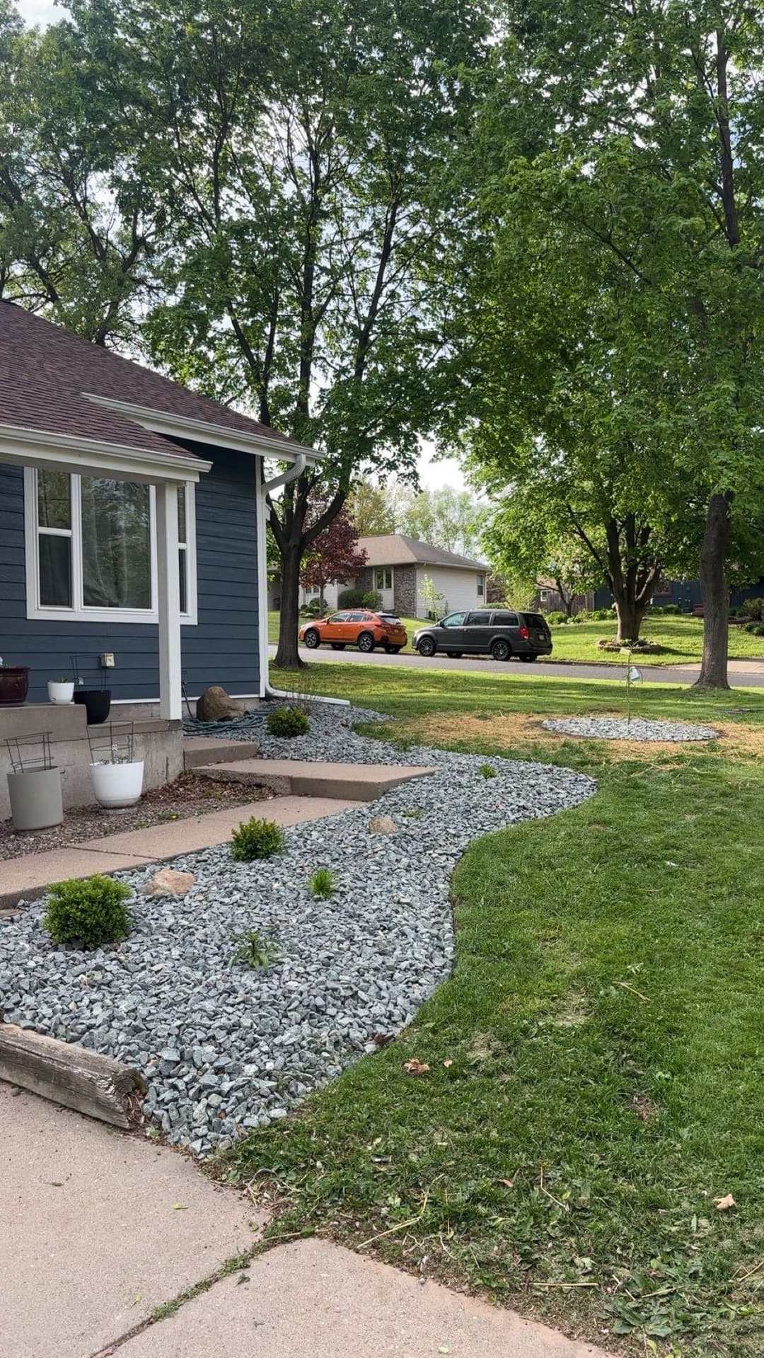 Blue house with a landscaped pathway and decorative stones, surrounded by green lawn and trees.